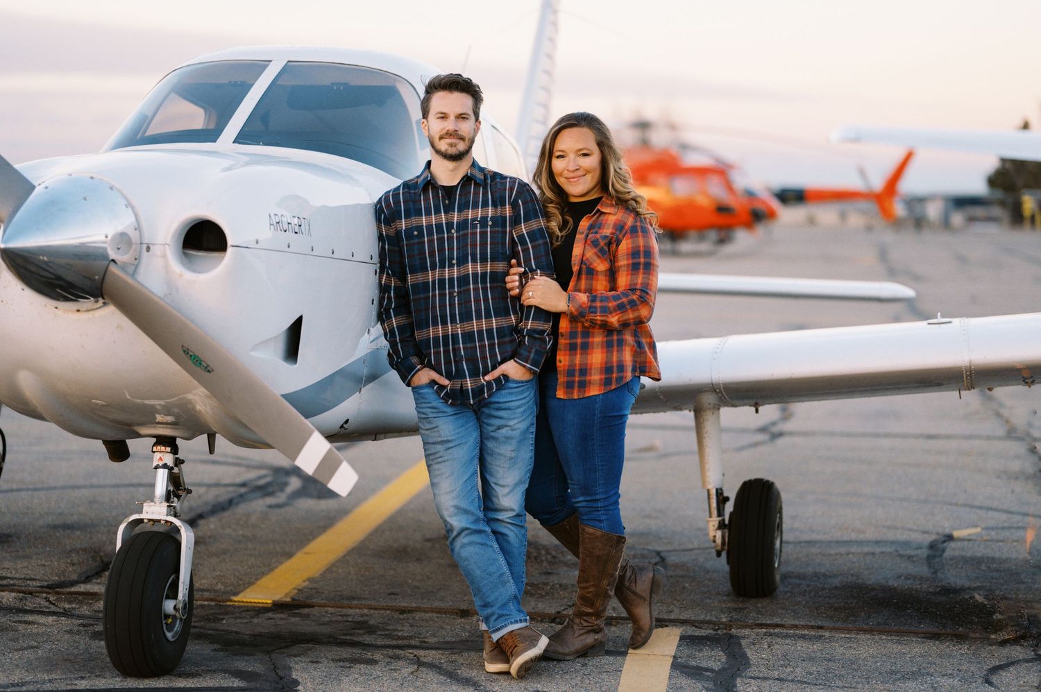 A couple in plaid shirts poses in front of a small private aircraft on the tarmac.