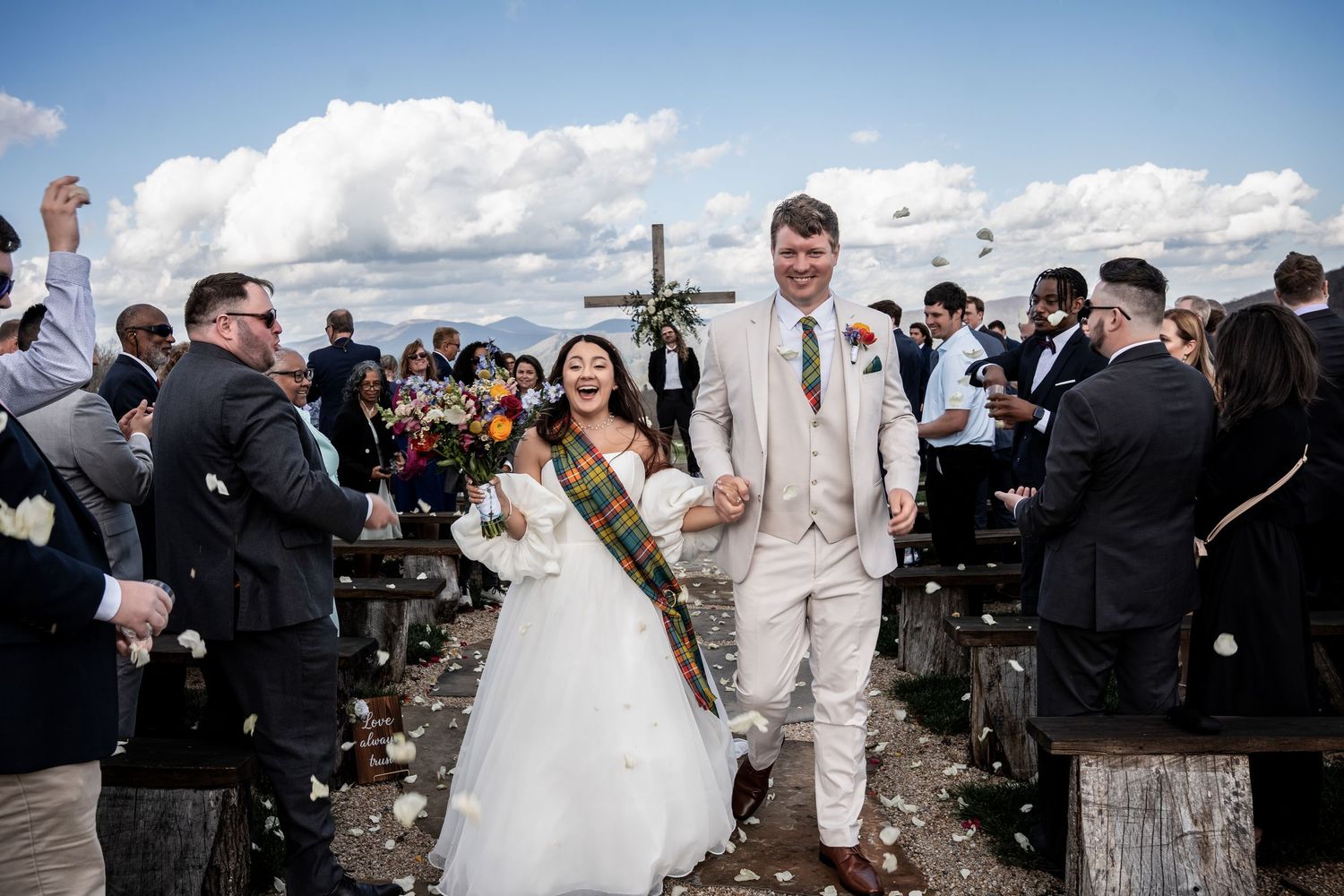 Couple walks down aisle at outdoor wedding ceremony while guests throw white flower petals on a sunny day with clouds.