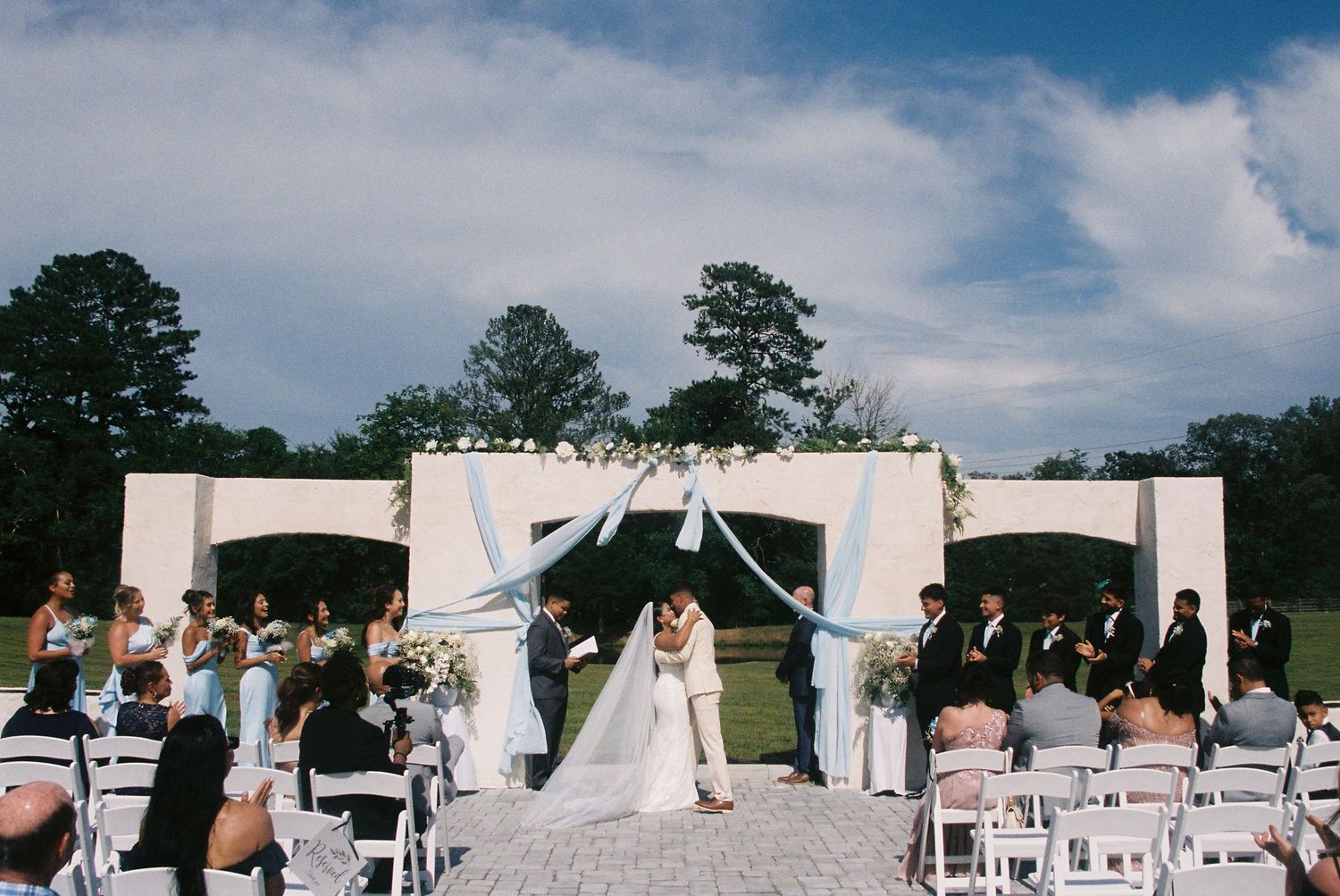 Elegant outdoor wedding ceremony setup with white draped archways and rows of chairs on a sunny day.