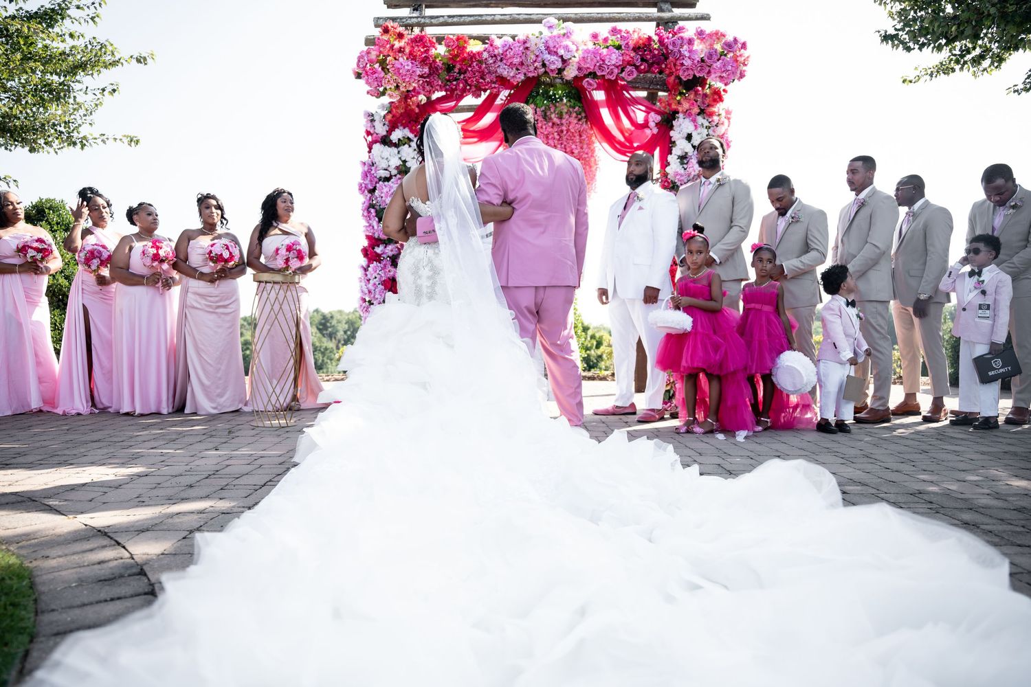 A wedding ceremony under a pink floral arch with bridesmaids in pink dresses and groomsmen in grey suits looking on.
