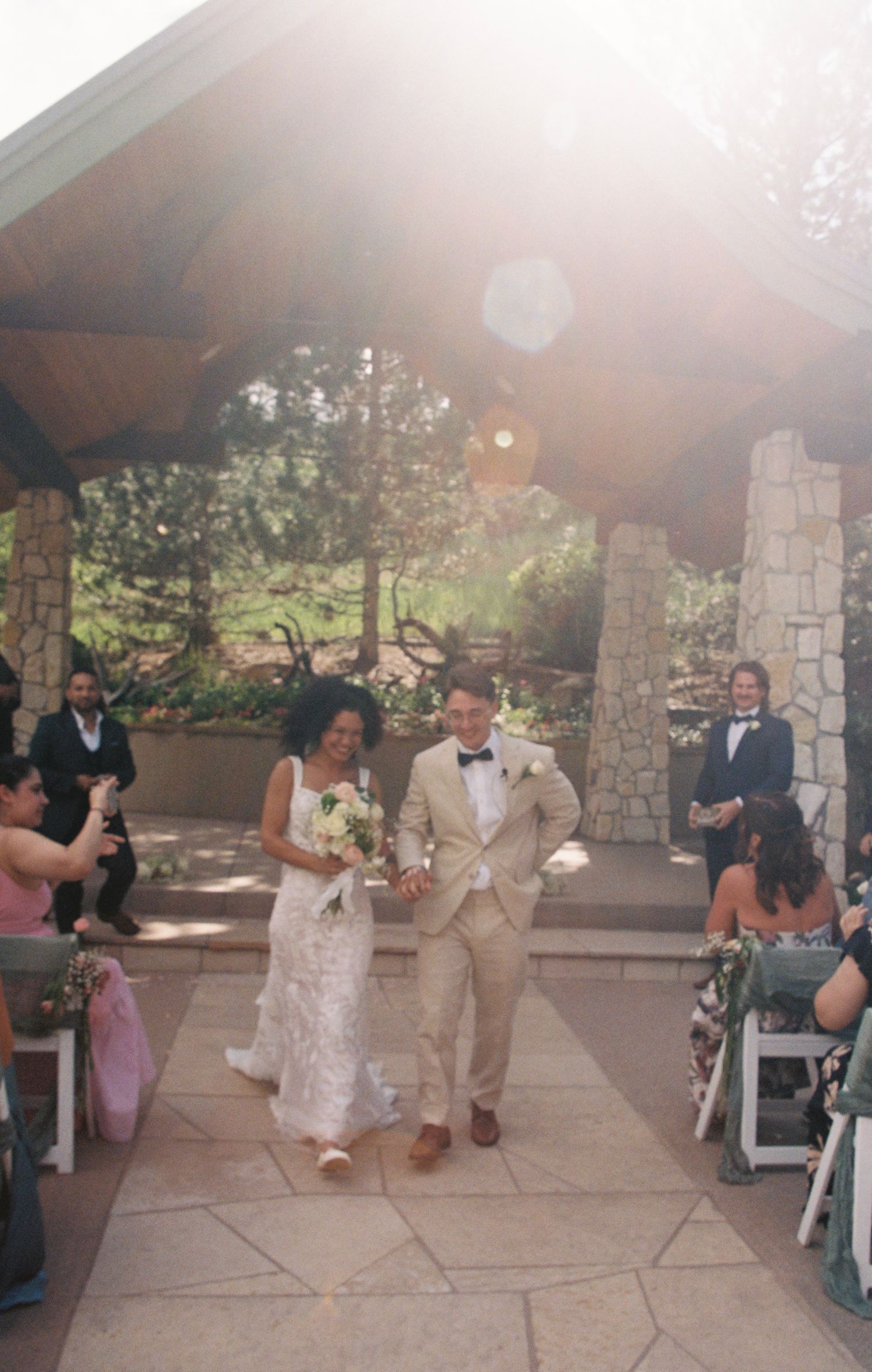 A happy couple walks down a stone-lined aisle at an outdoor wedding ceremony under a covered pavilion on a sunny day.