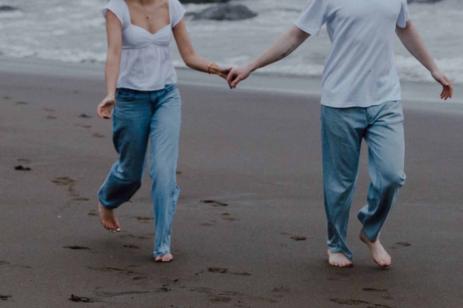 Two people holding hands while walking barefoot along a misty beach in casual jeans and tops.