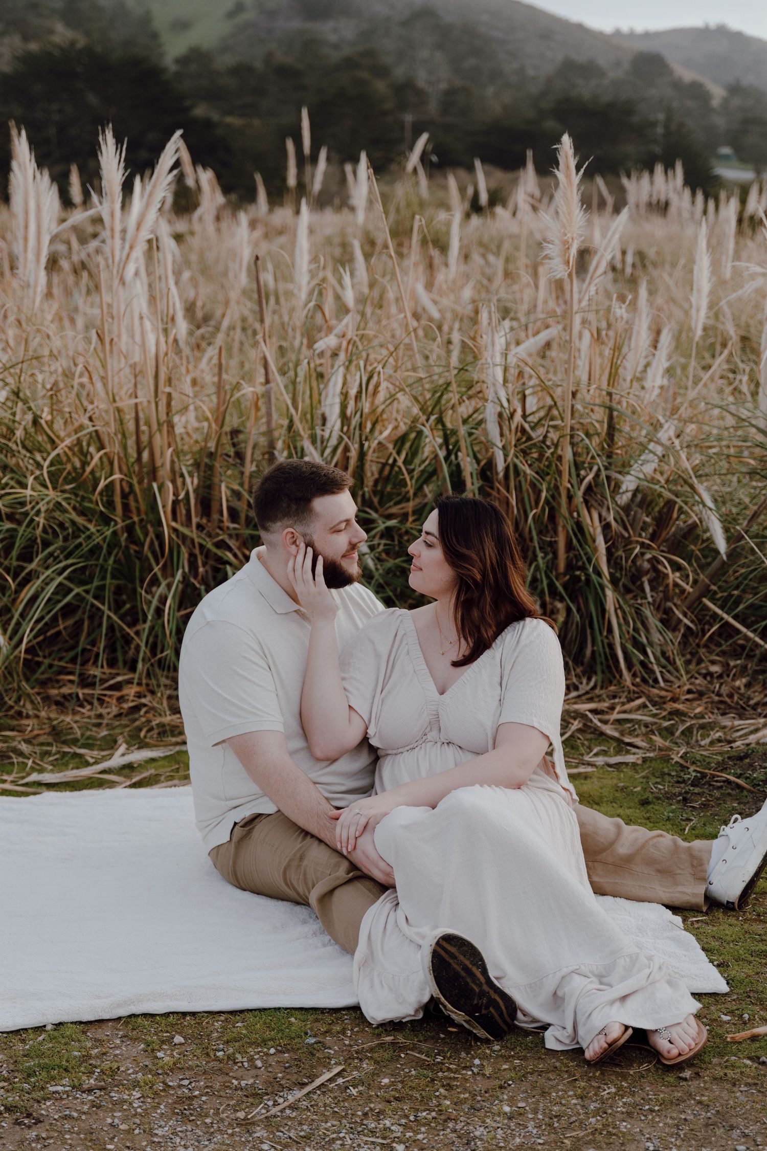 Romantic couple in white outfits sitting on blanket against tall pampas grass during outdoor photo session.