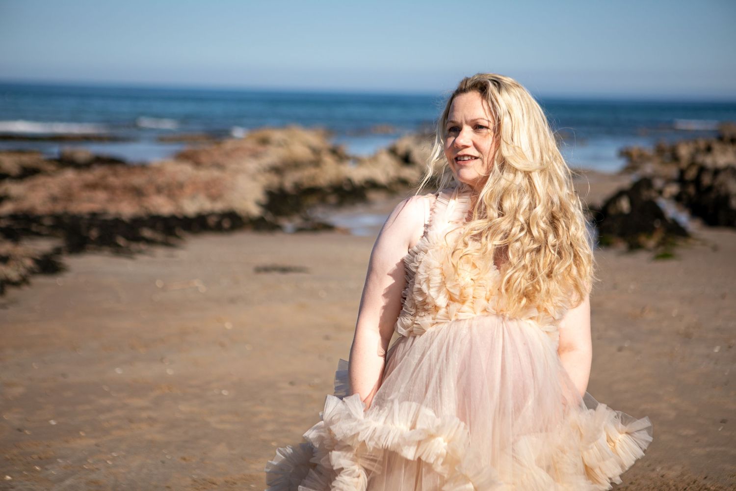 A person with long blonde hair wearing a beige tulle dress stands on a rocky beach with ocean waves in the background.