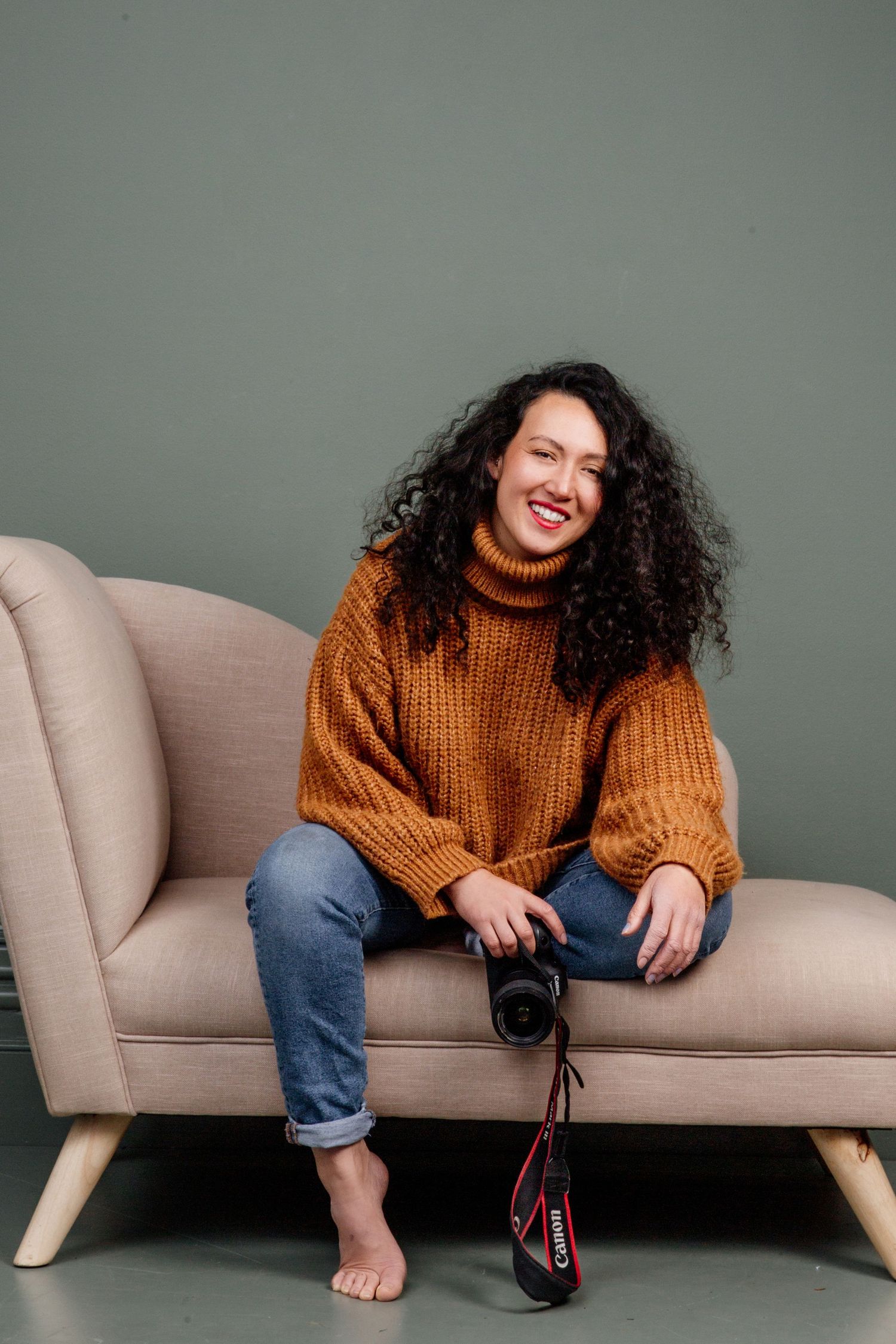 A person in a brown knit turtleneck sweater smiles warmly with curly dark hair against a light neutral background.