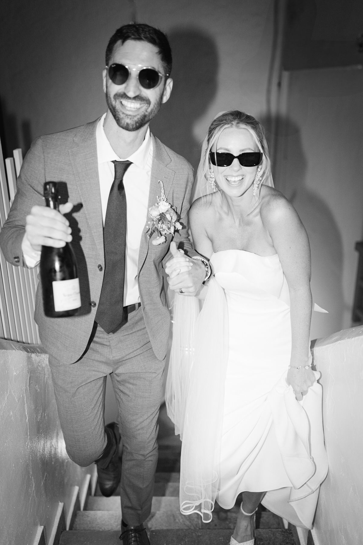 A joyful bride and groom wearing sunglasses walk together on stairs while holding a champagne bottle in black and white. Vogue.