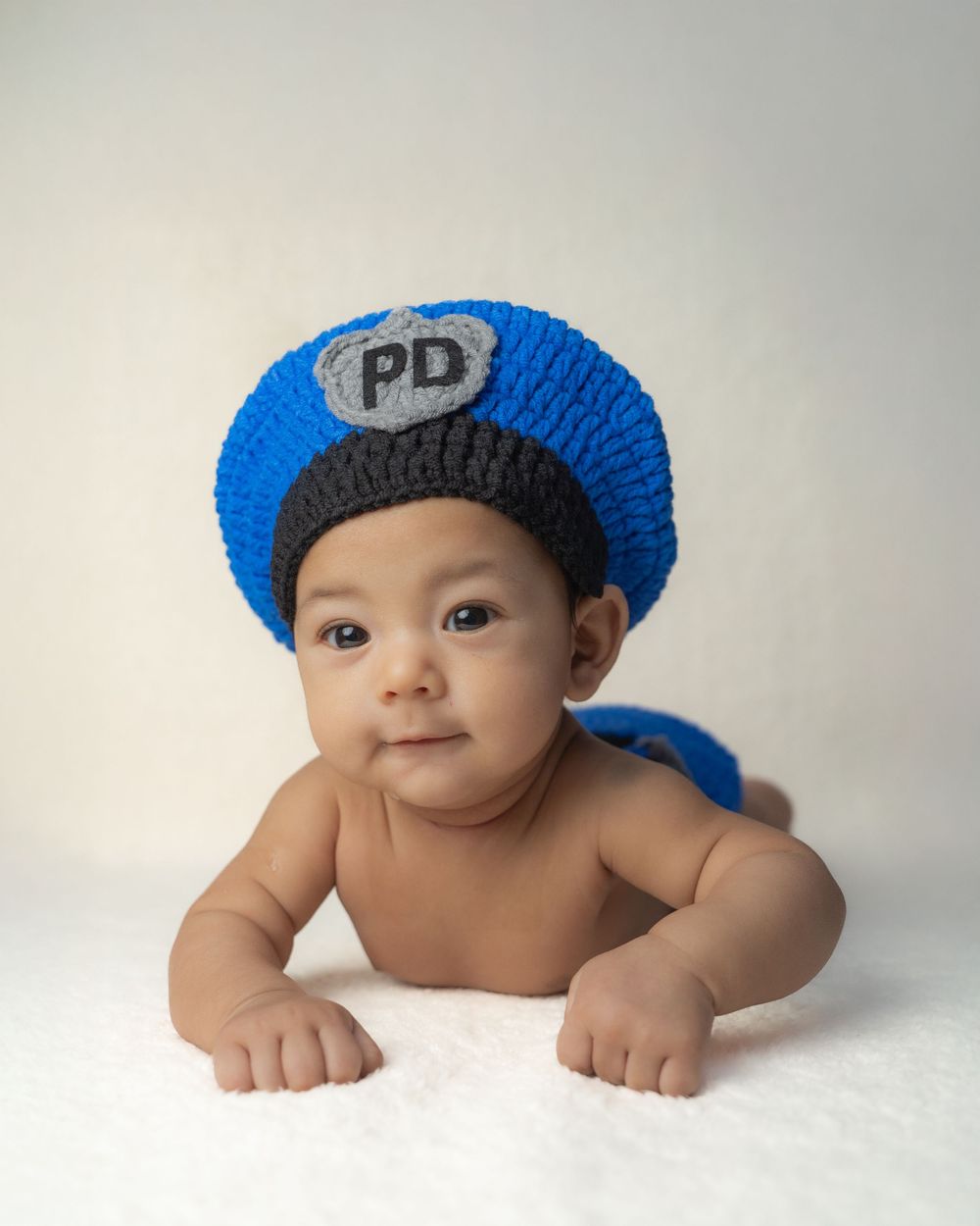 Baby wearing a bright blue knit police hat lies on white backdrop during professional photo shoot.