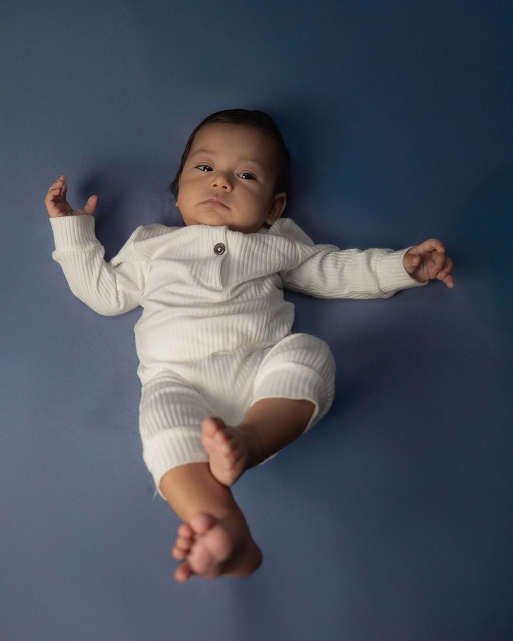A baby wearing a white ribbed sweater outfit lies on a blue background.