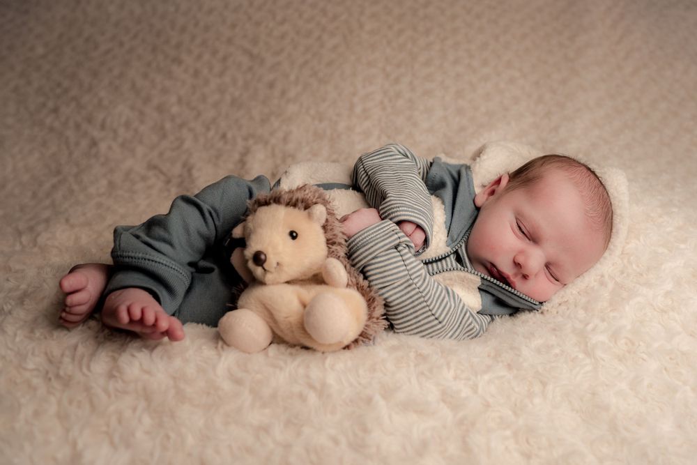 Baby sleeping peacefully on cream blanket while cuddling plush teddy bear and wearing gray outfit.