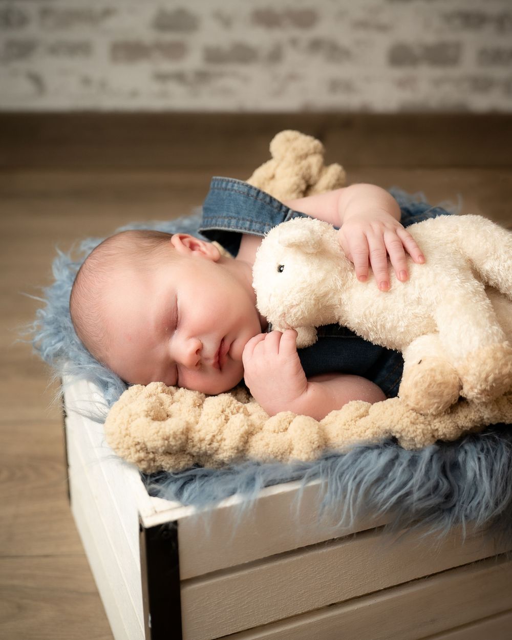 Sleeping infant in blue outfit cuddled up with soft beige teddy bear on cozy fur-lined prop bed.
