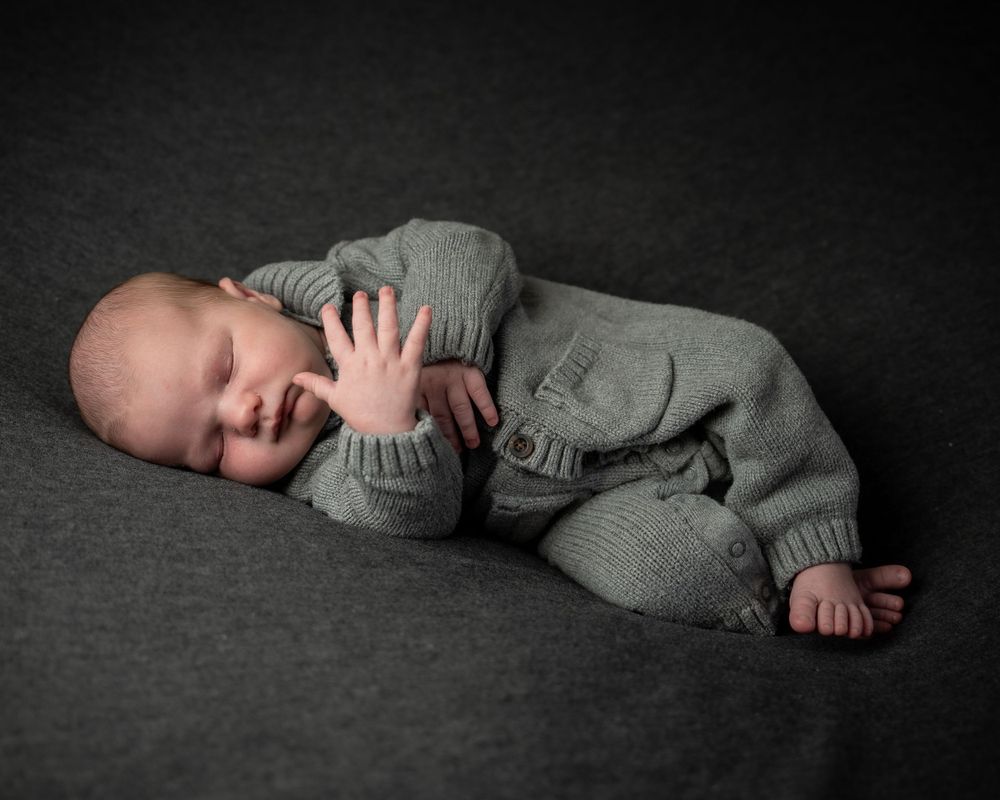 A sleeping baby wearing gray knit clothing lies on a dark textured backdrop in a professional studio setting.