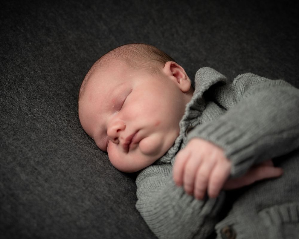 Sleeping newborn in gray sweater rests peacefully on dark gray backdrop in professional portrait.