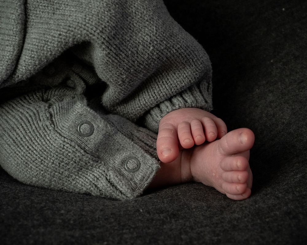 Tiny baby feet peek out from underneath a cozy gray knit sweater on a dark background.
