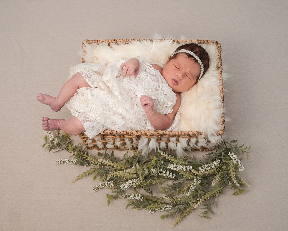 A sleeping newborn in a white lace dress lies on soft white fur with evergreen branches decorating the bottom of the frame.