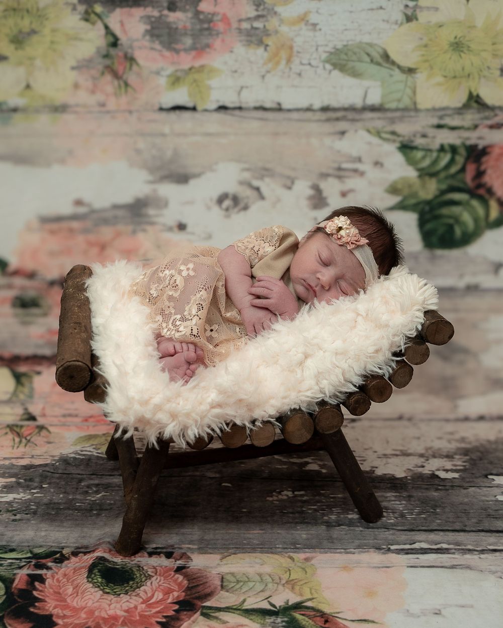 Sleeping newborn rests on wooden vintage bench covered in fluffy white fur with floral backdrop.