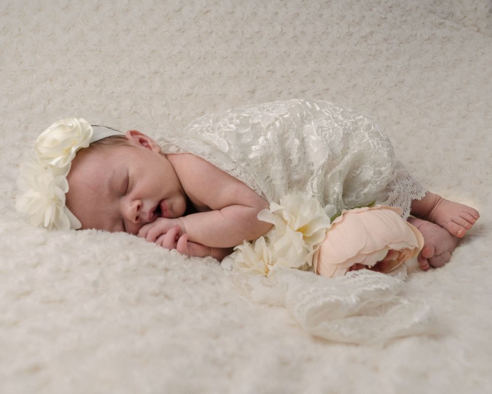 Sleeping newborn in cream-colored lace outfit resting peacefully on soft white textured blanket.