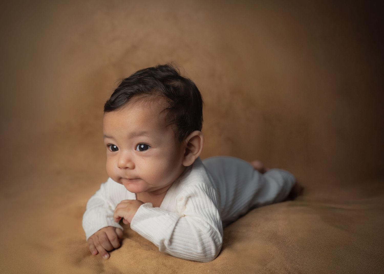 Baby wearing white sweater lies on stomach against brown backdrop during professional portrait session.