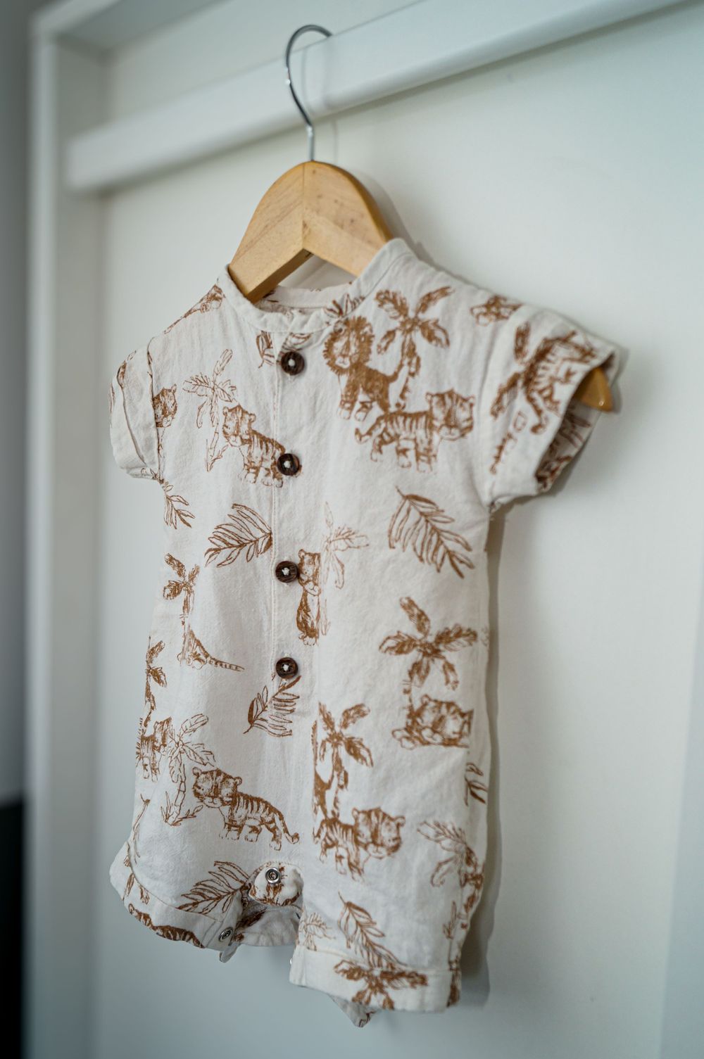 A baby romper with brown safari animal print patterns hangs on a wooden hanger against a white wall.