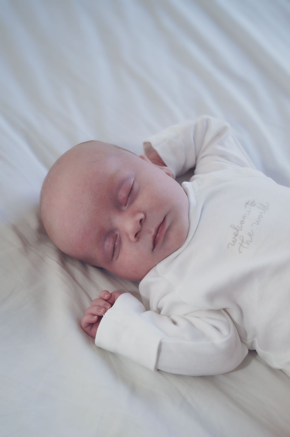 A sleeping baby wearing a white sleeper lies peacefully on white bedding.