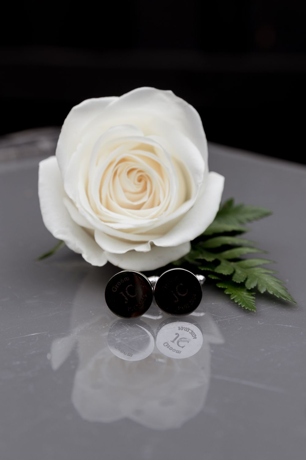 A white rose and cufflinks arranged on a reflective surface for a wedding detail shot.