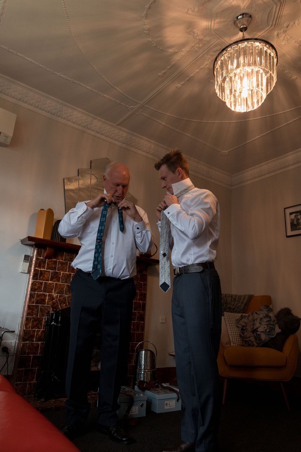 Two people adjusting their formal attire beneath a crystal chandelier in an elegant room.
