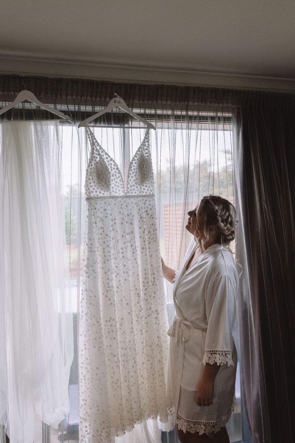 Elegant white lace wedding dress hanging by window as figure in robe admires it in morning light.