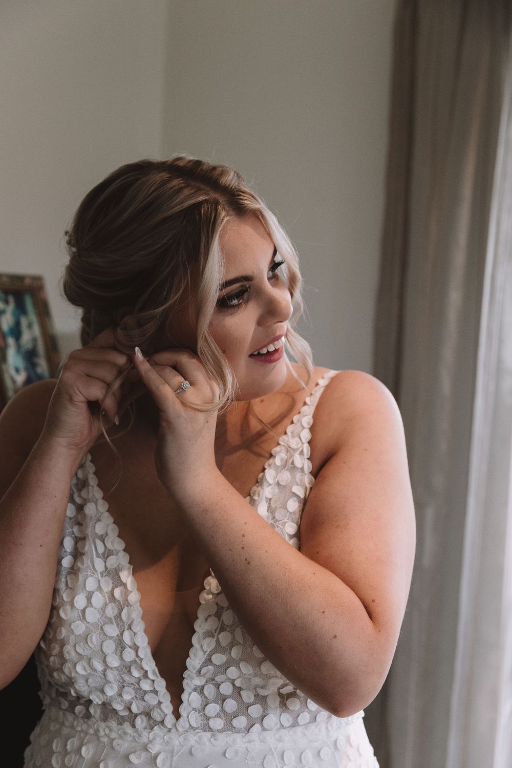 Close up of bride putting on earrings while wearing intricate white beaded wedding gown.