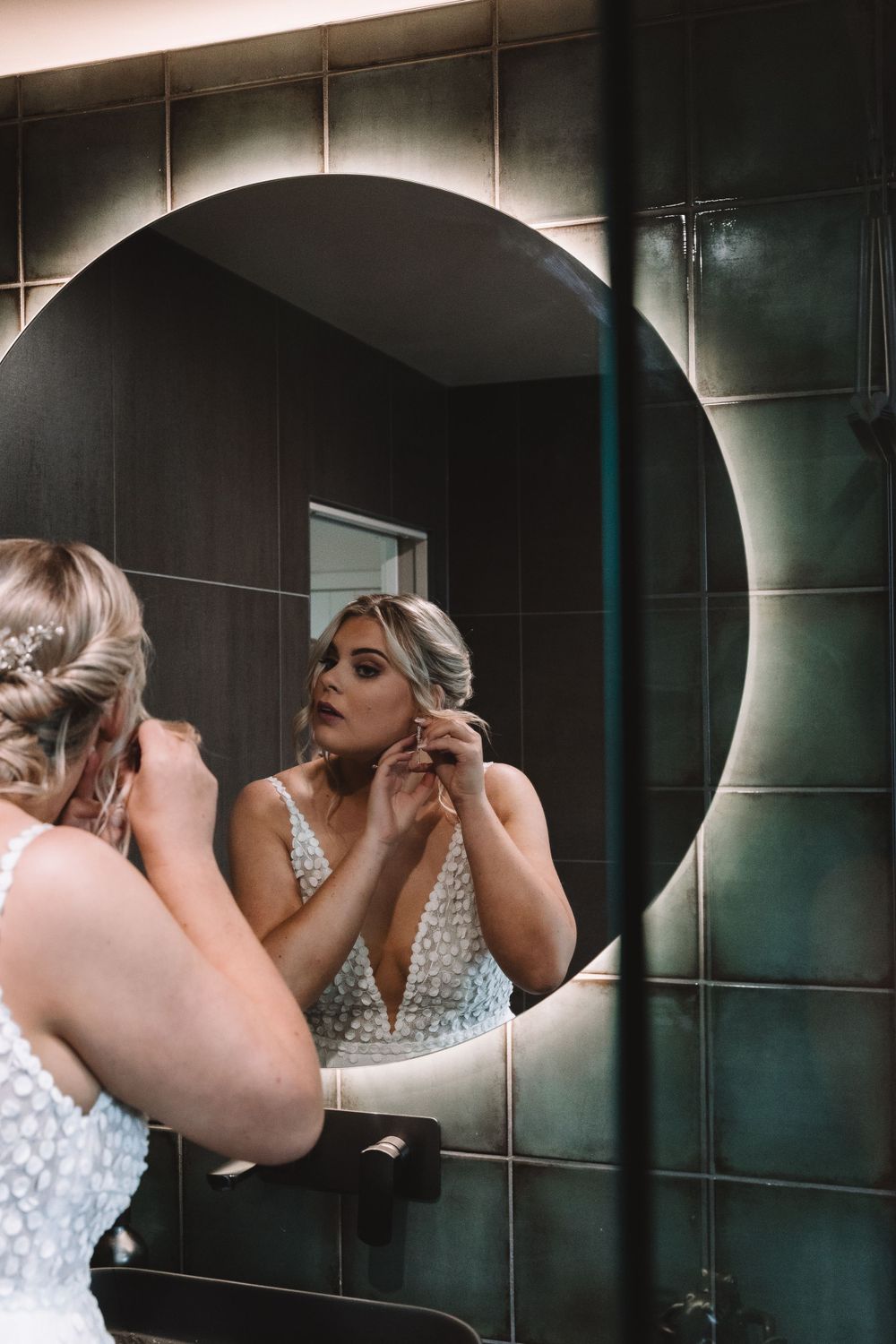 Artistic mirror reflection shot of bride putting on jewelry in modern bathroom.