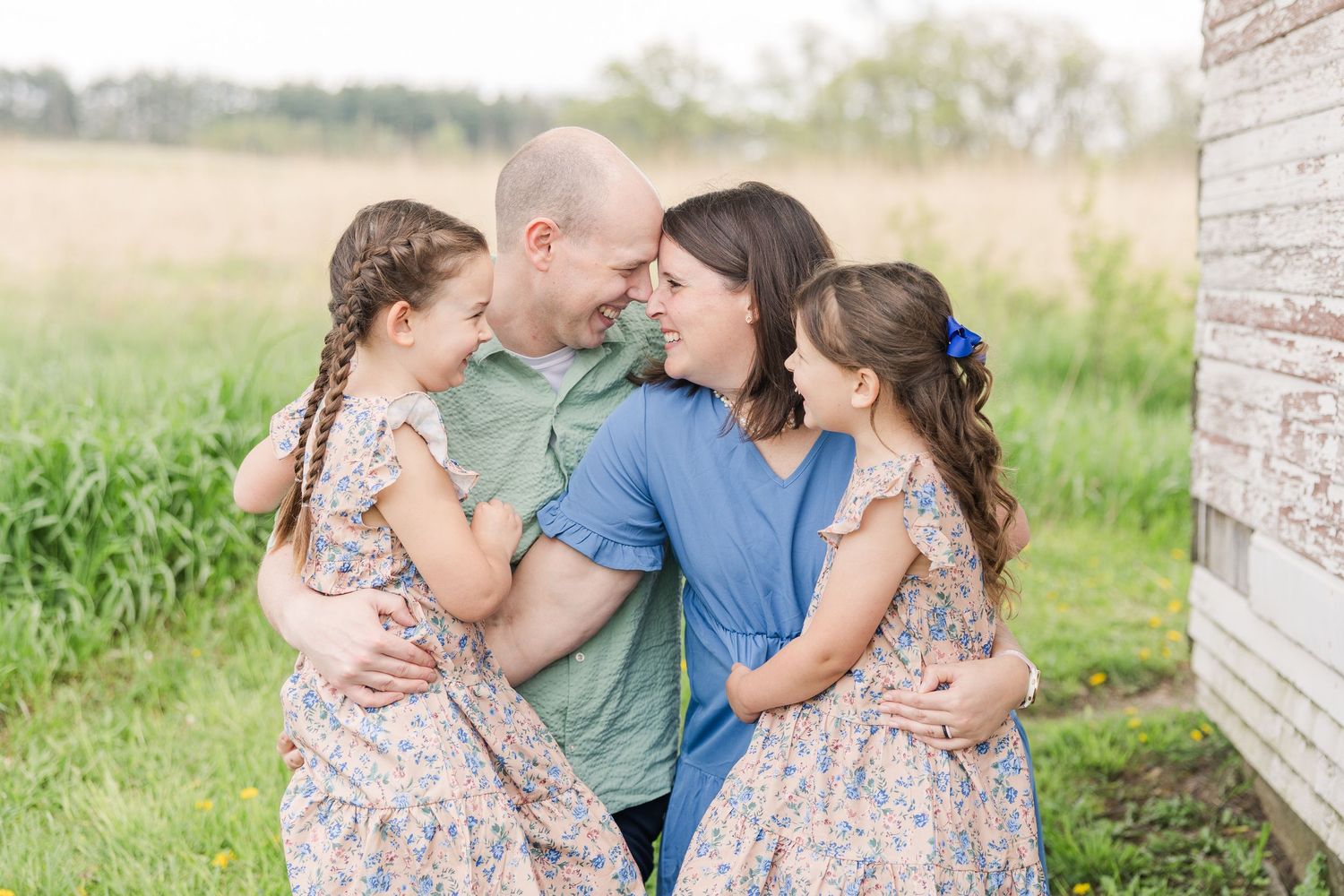 A family embraces outdoors in matching floral dresses against a rustic barn wall and green field.