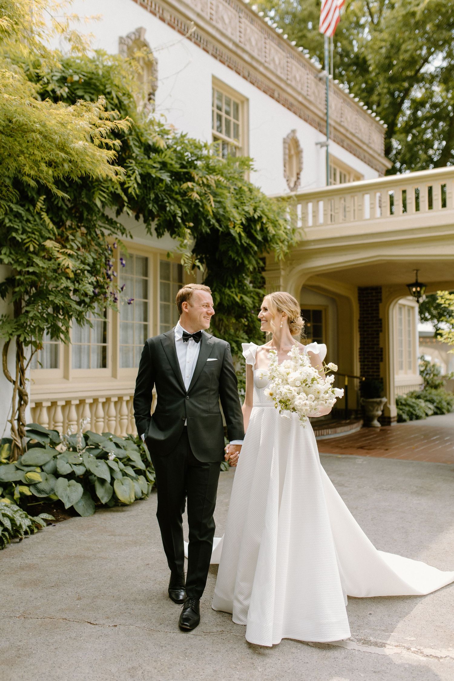 A newlywed couple walking together outside an elegant white mansion with classic architectural details and lush greenery.