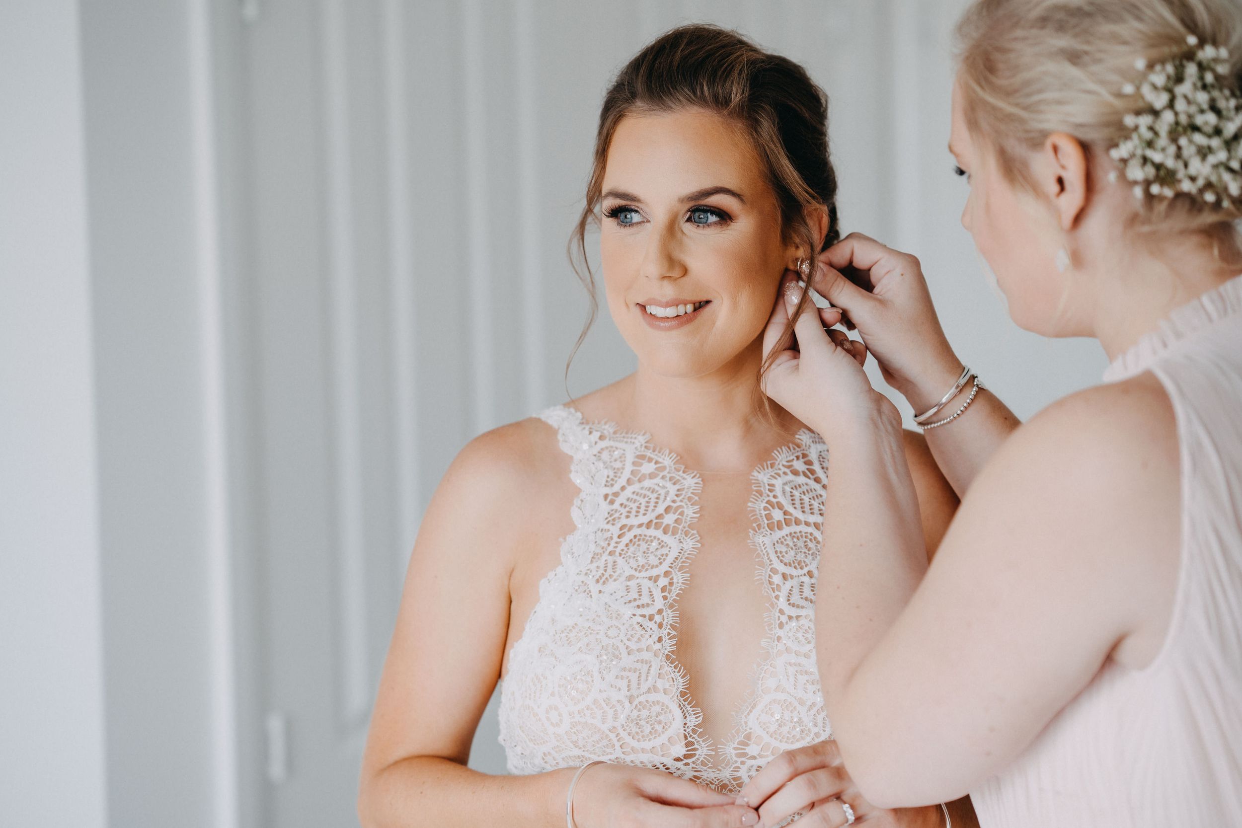 Two people getting ready for a wedding, one helping the other with jewelry while wearing a delicate lace gown.