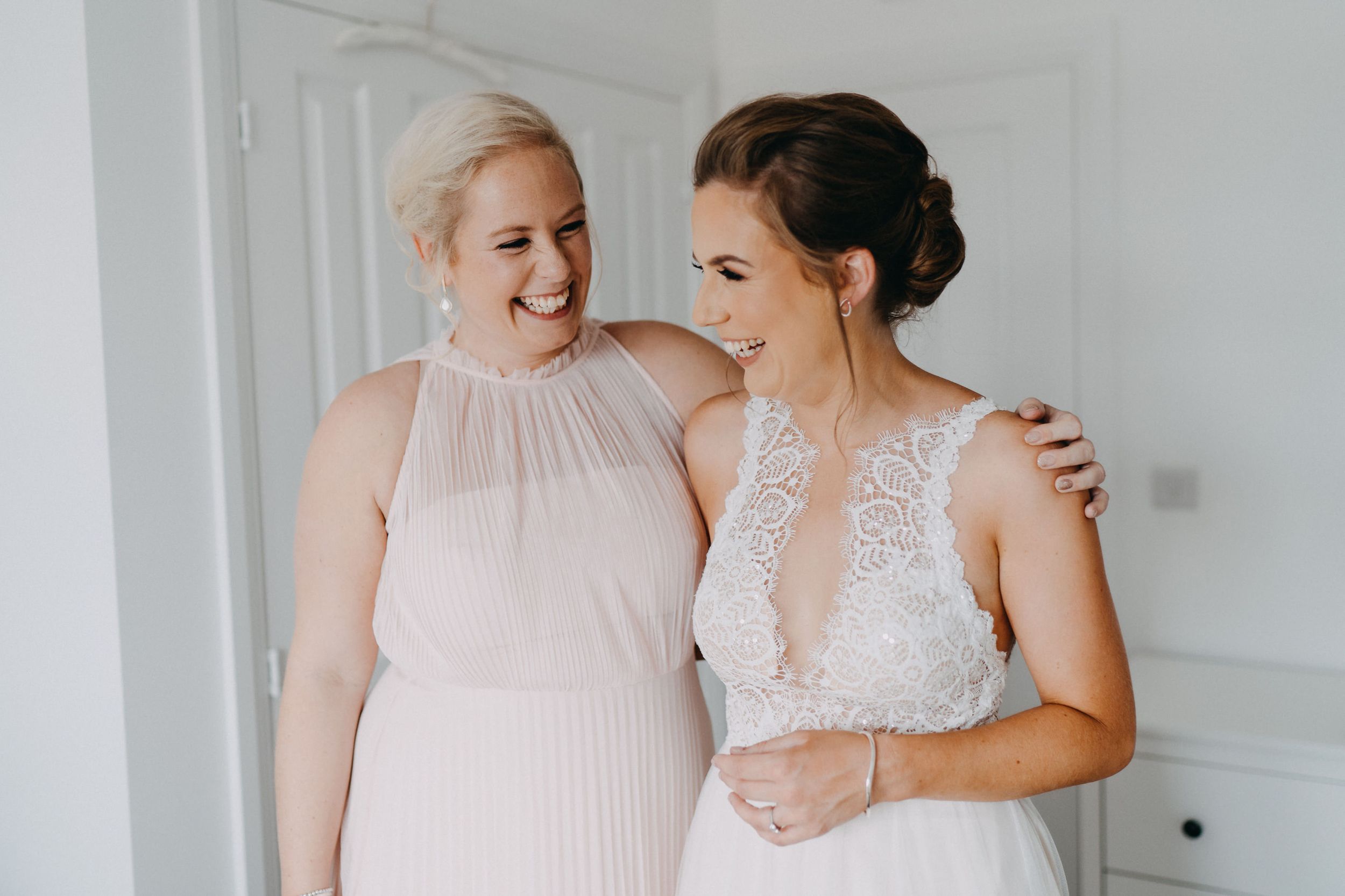 Two women in light pink and white dresses share a joyful moment together in front of a white wall.
