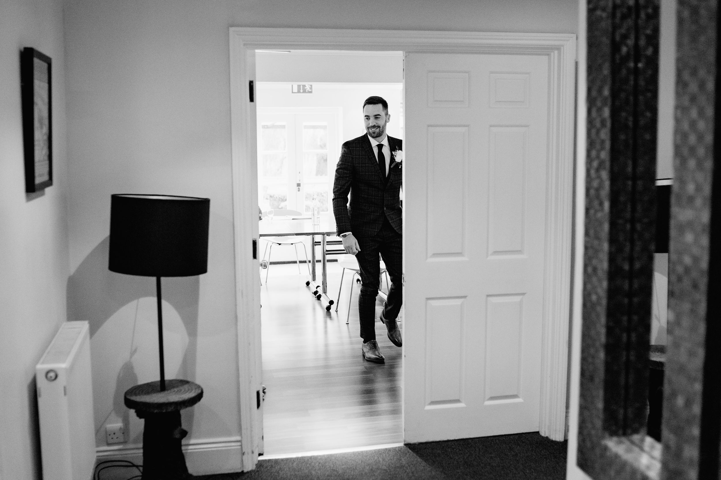 A silhouette stands in a doorframe wearing a dark suit against a bright background in black and white.