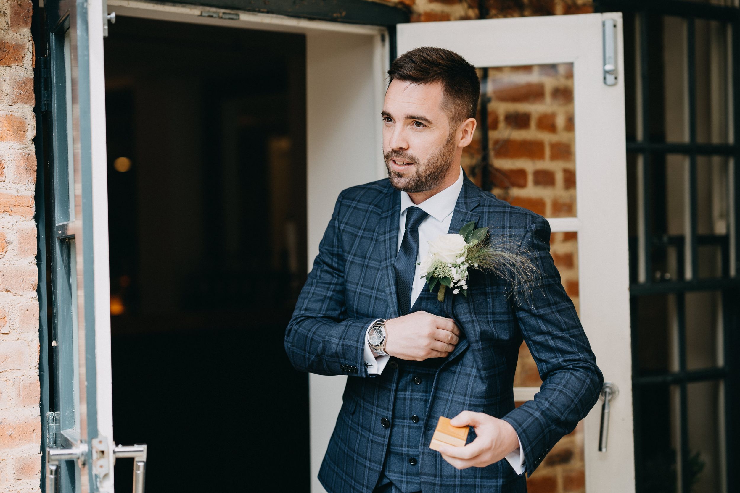 A person in a tailored navy windowpane suit jacket with boutonniere adjusts their sleeve by a brick wall.