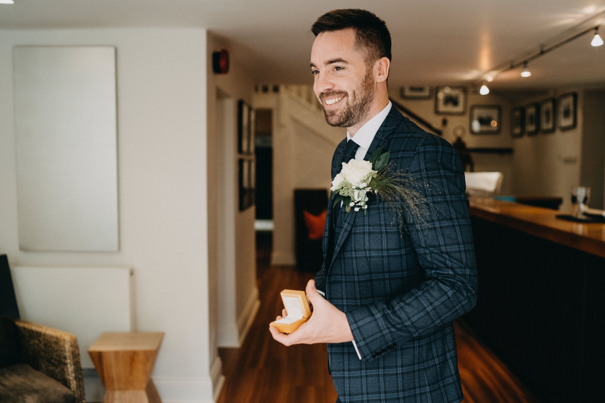 Groom wearing navy checkered suit with boutonniere smiles while holding coffee cup in indoor venue.