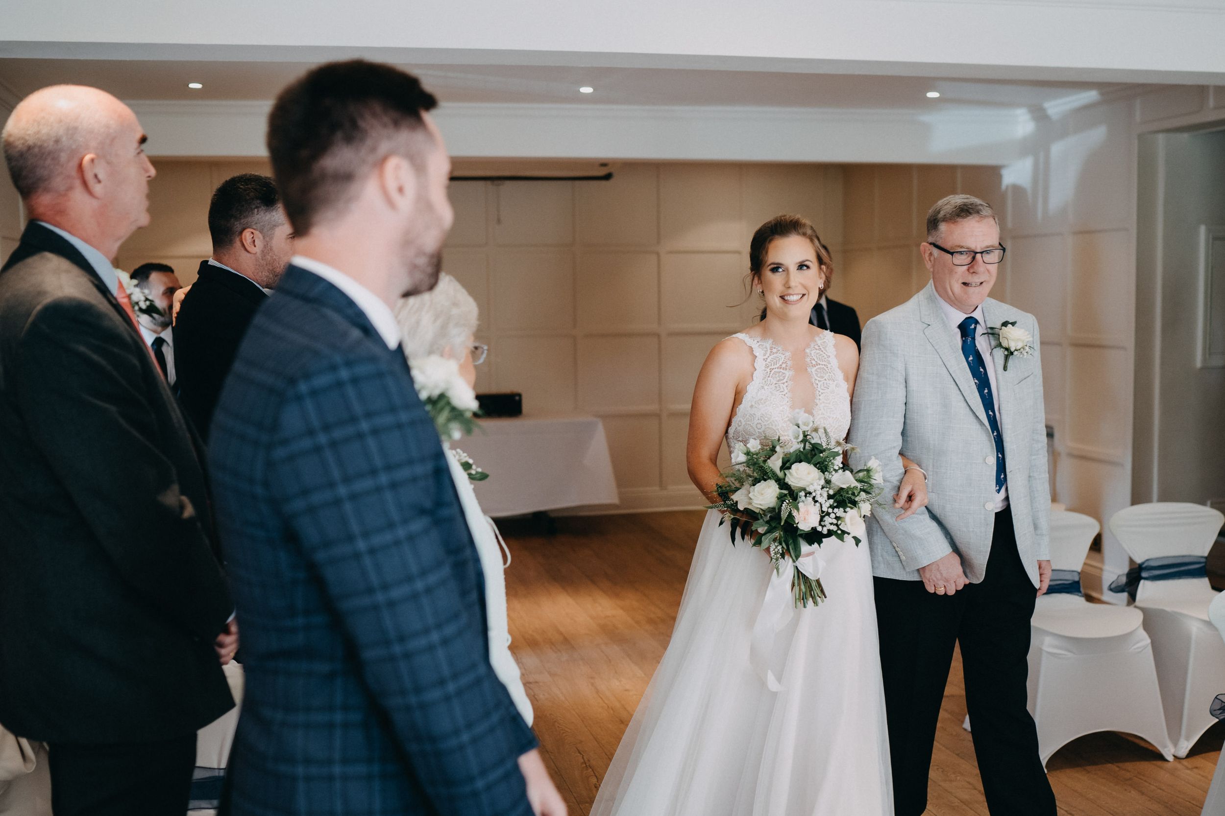 A wedding couple stands in an indoor venue while guests in suits gather around during a ceremony.