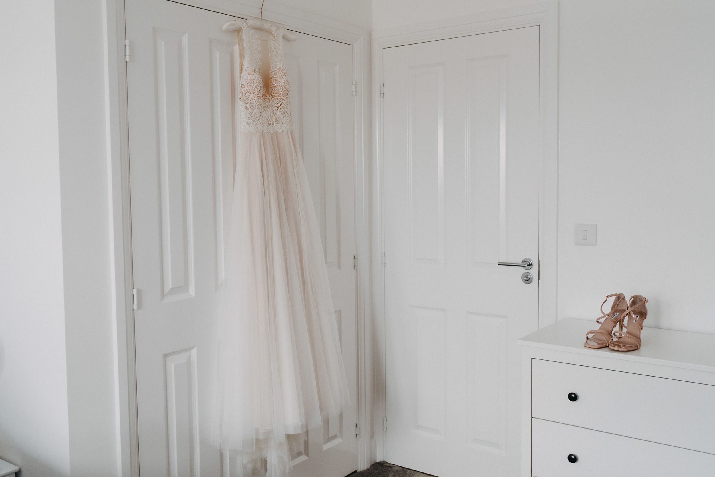 A light blush bridal gown hangs elegantly on a white wardrobe door beside a dresser with rose gold shoes.