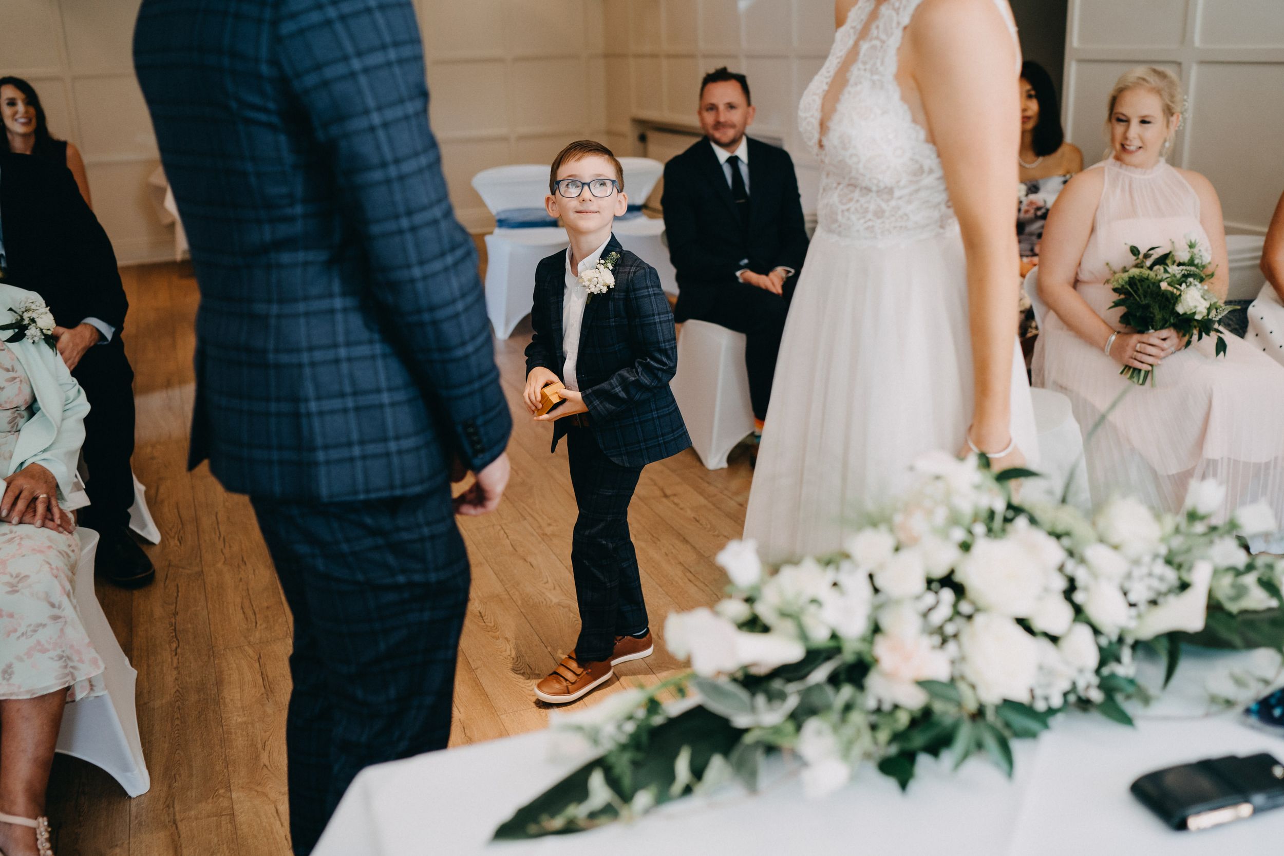 Wedding ceremony taking place in a light-filled room with white floral arrangements and guests seated in rows.