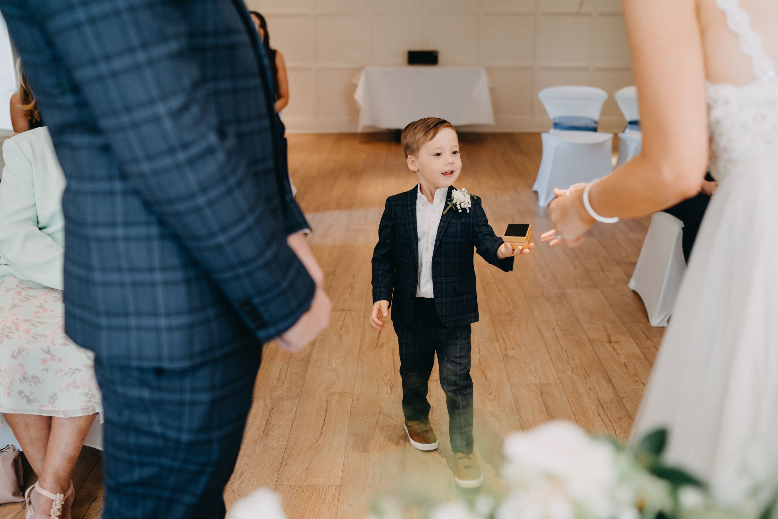 A small child in a formal black suit stands between adults during a wedding ceremony on hardwood floors.