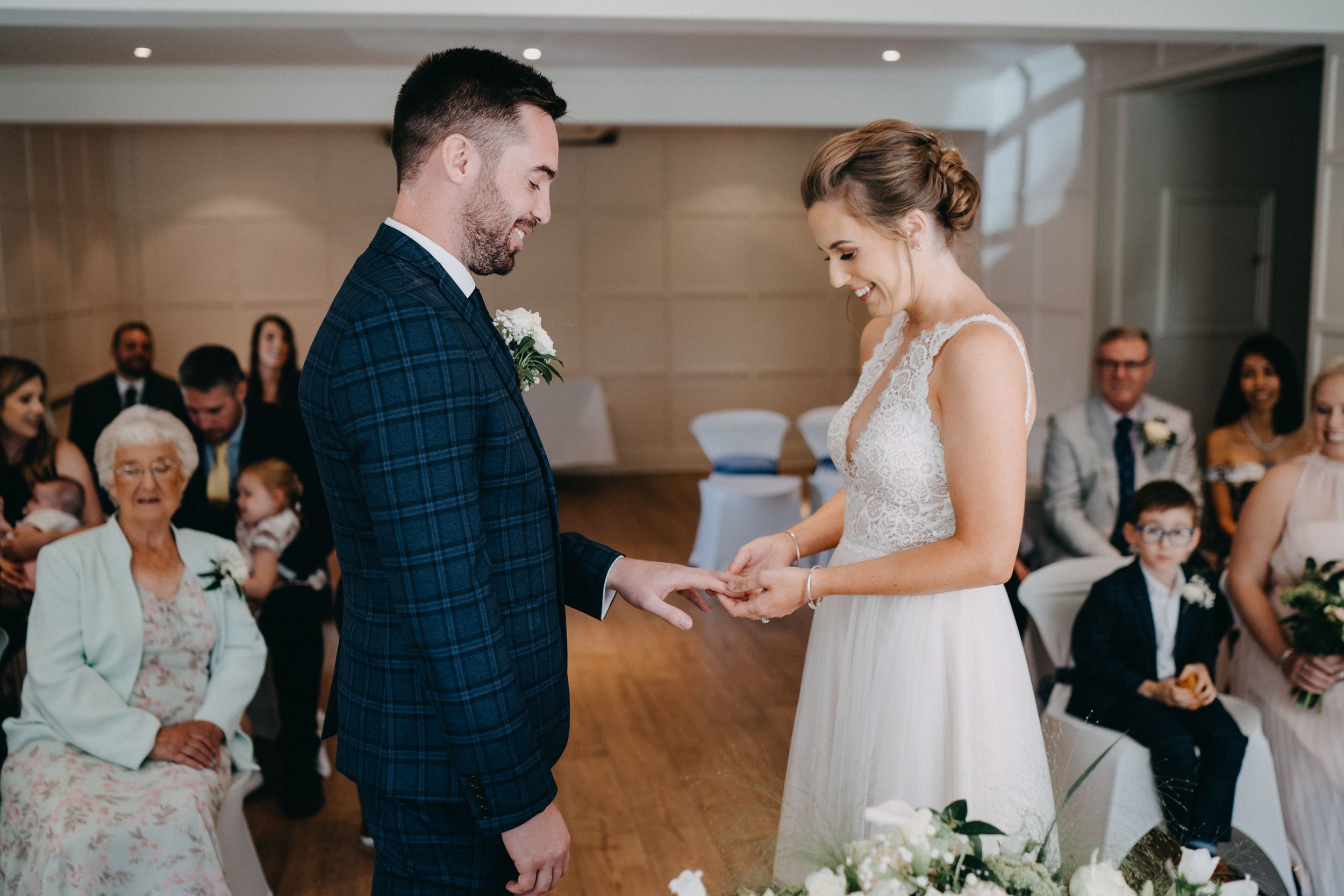 Bride and groom exchange rings during an intimate indoor wedding ceremony as guests look on in the background.