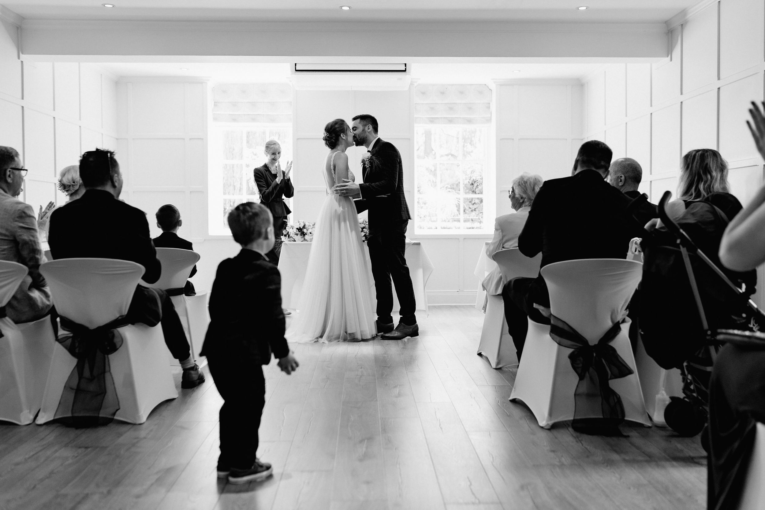 Black and white photo of indoor wedding ceremony at The Old Hall with guests watch from white chairs in bright room.