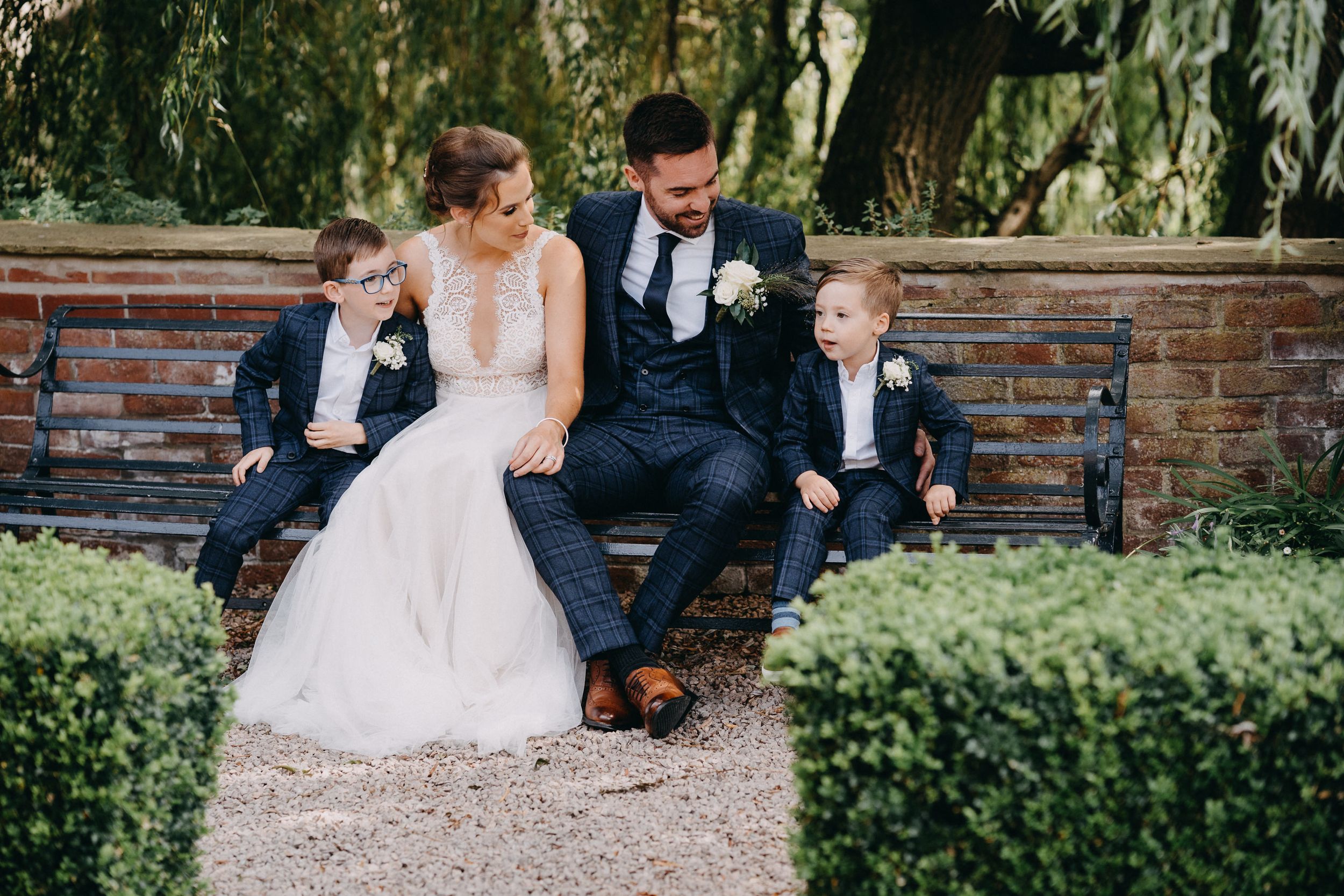 Wedding party relaxes on a rustic brick wall surrounded by lush greenery at an outdoor garden venue.