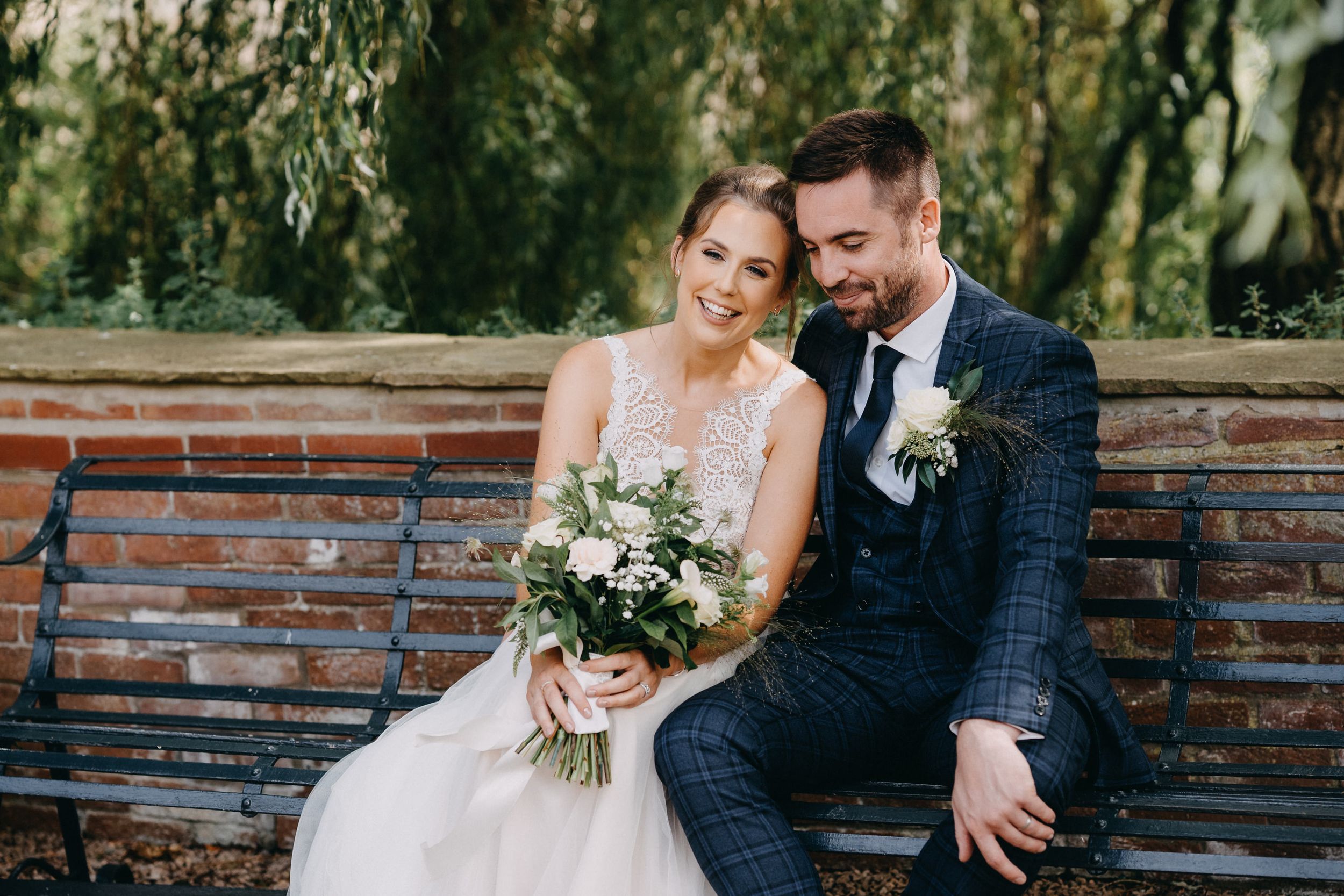 A couple sits together on a brick wall beneath weeping willow trees holding a white floral bouquet.