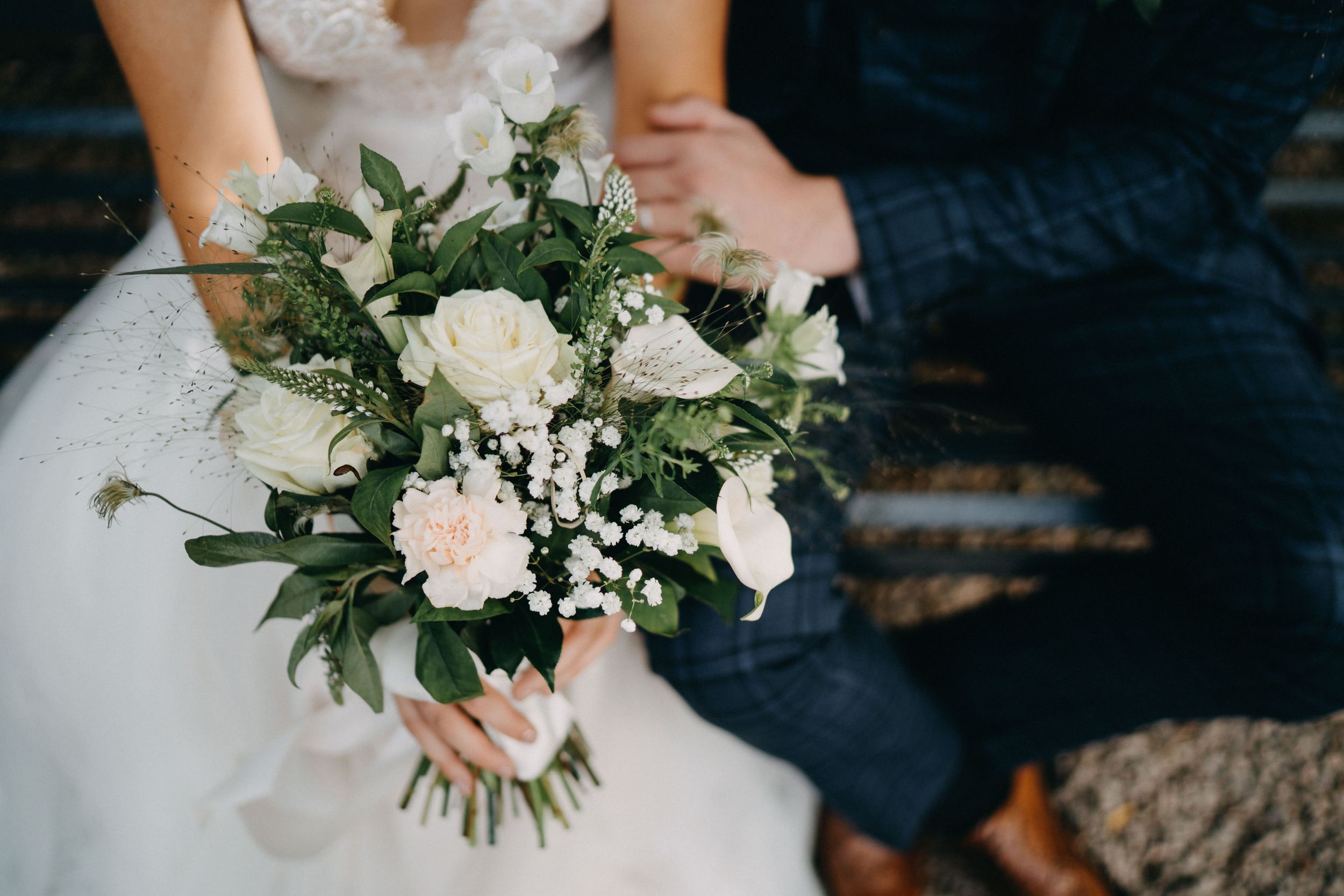 Close-up of elegant white and blush wedding bouquet with roses and greenery held against bride's dress.