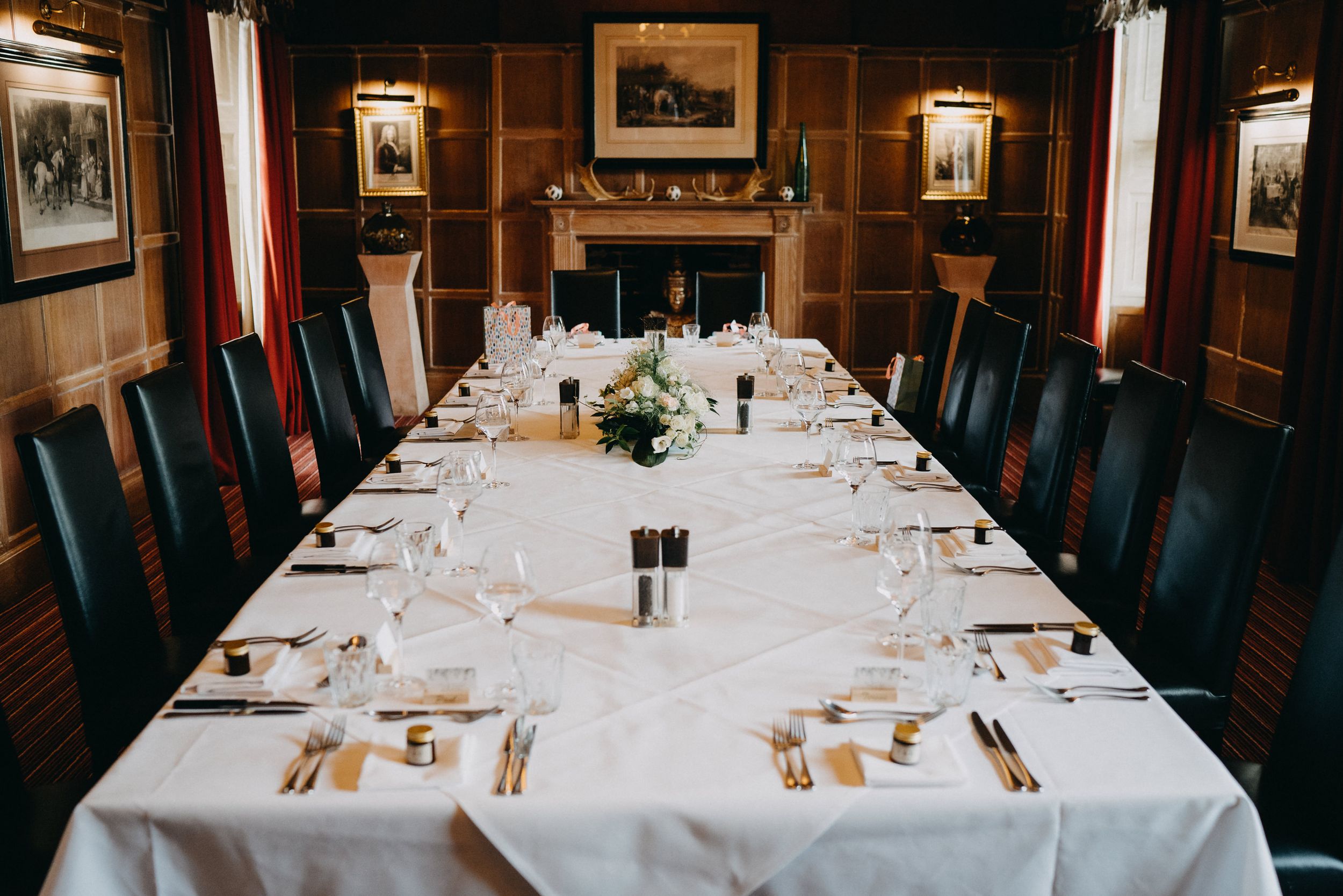 Elegant formal dining table set with white linens and place settings in a wood-paneled room with a fireplace.