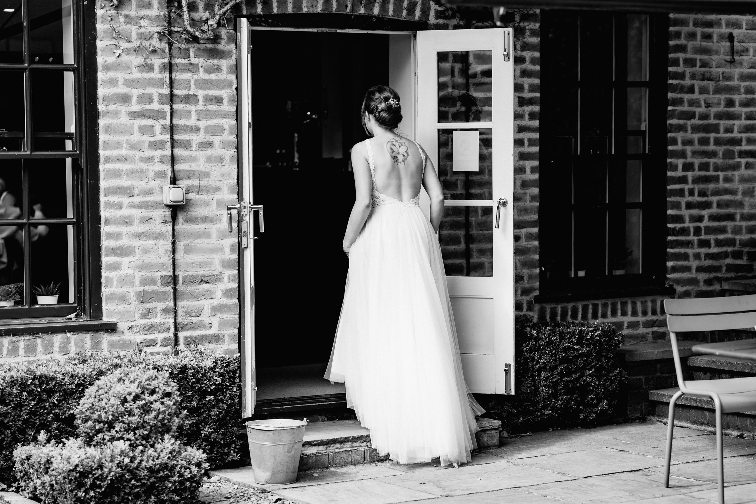 Black and white photo shows someone in a flowing white wedding dress at an entrance to a brick building.