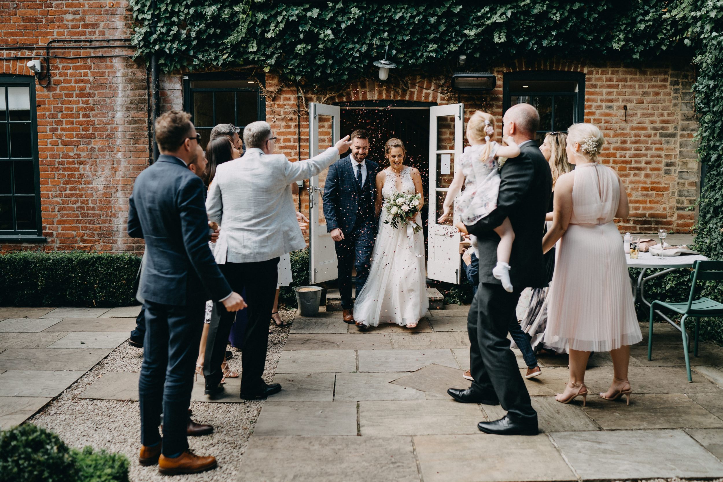 Wedding party gathers on stone patio outside rustic brick building with ivy during intimate celebration.