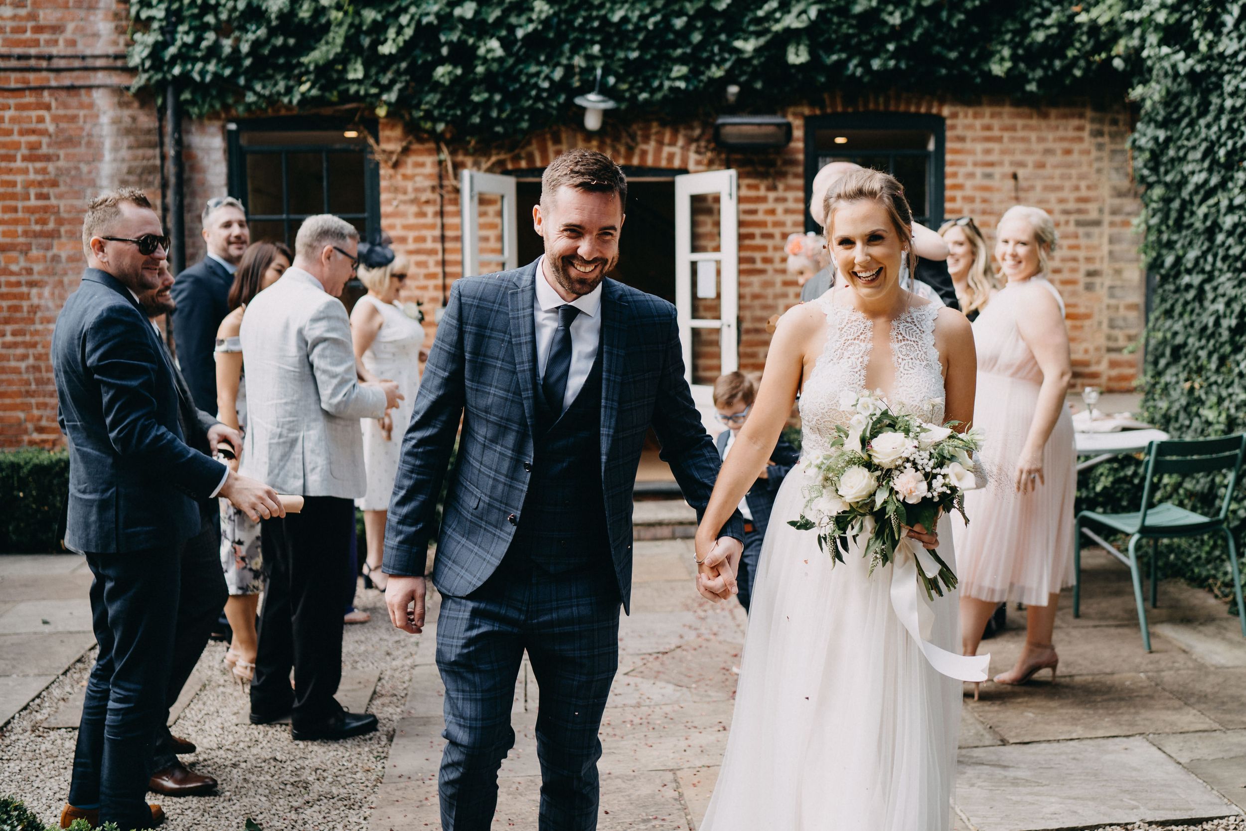 Wedding party gathers outside brick venue with ivy as bride holds white floral bouquet in lace gown.