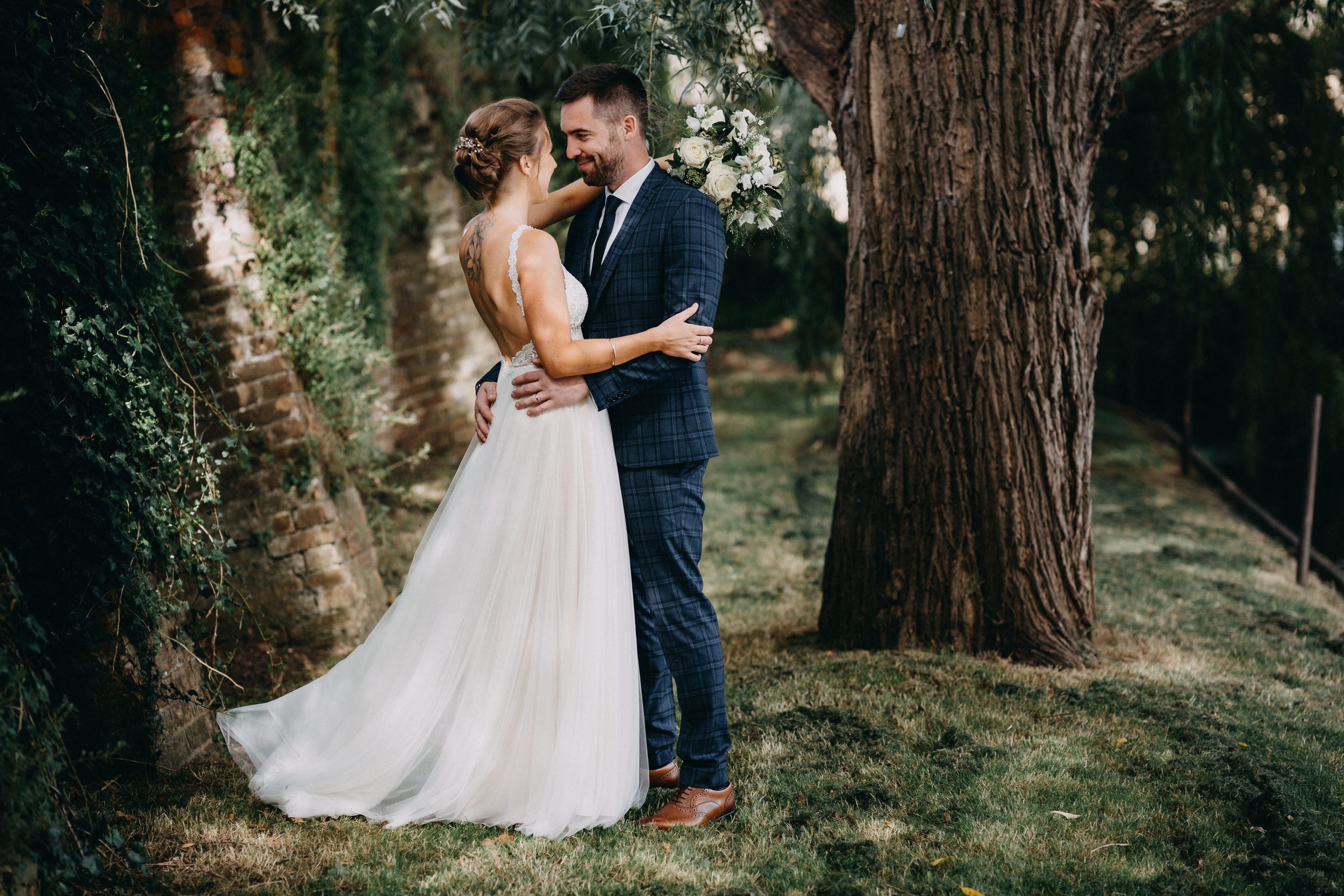 A romantic couple embraces in a sun-dappled forest setting while wearing wedding attire.