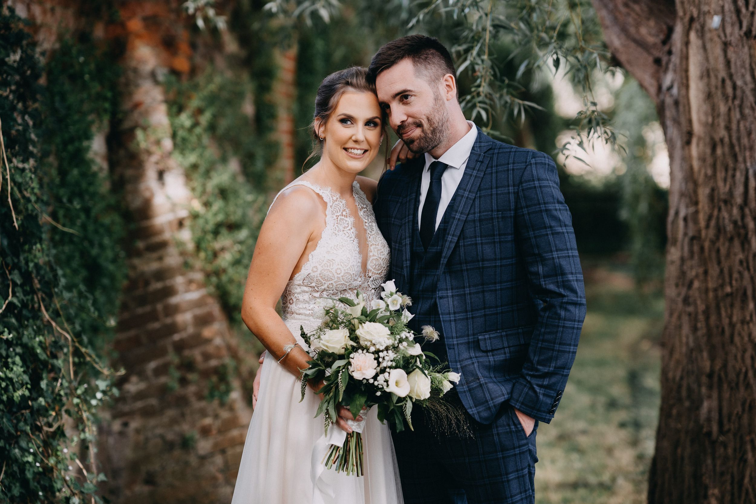 A couple shares an intimate moment in a forest setting with the bride holding a white floral bouquet.